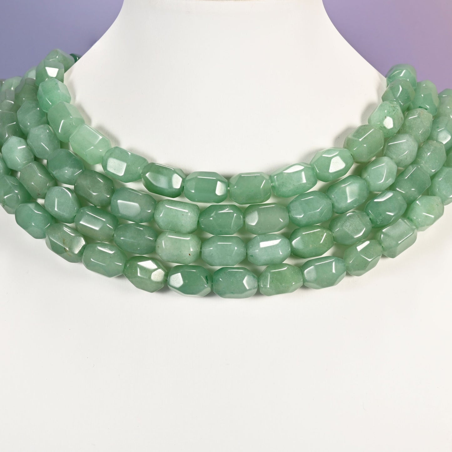 Multi-strand necklace of green faceted Aventurine barrel beads laid on a white mannequin neck form against a lavender background.