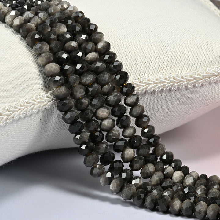 Silver obsidian faceted rondelle beads in black and smoky gray with a silver sheen, several strands draped over a cream cushion with braided trim