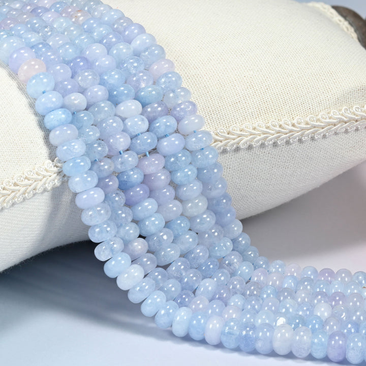 Aquamarine Blue dyed jade gemstone rondelle beads draped over a cushion in soft rows, a light aquamarine blue with a scatter of light purple beads mixed through the strand