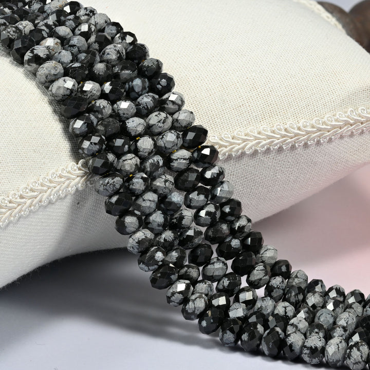 Snowflake jasper faceted rondelle beads 5x8mm in glossy black with gray snowflake patches, several strands draped over a white cushion
