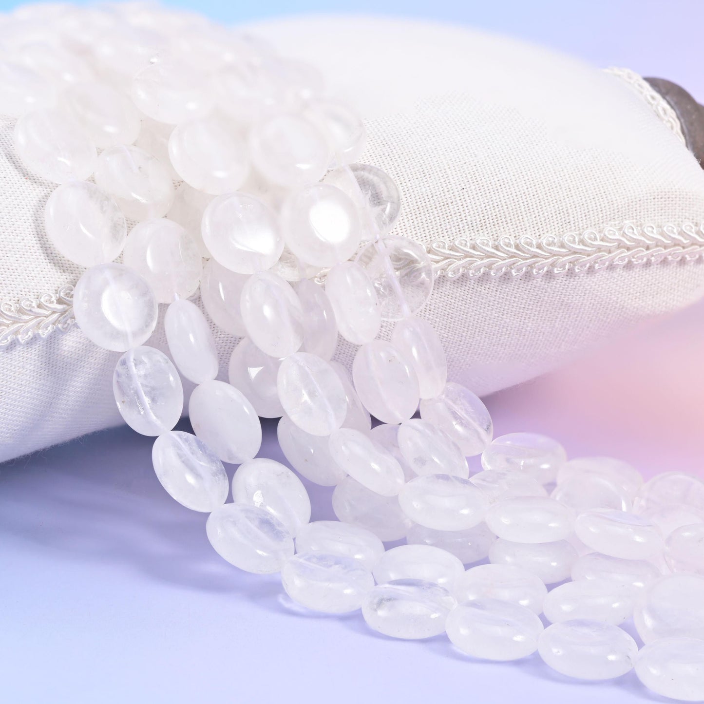 Clear quartz coin strands resting on a cream linen cushion, frosted white discs on a pale lilac backdrop