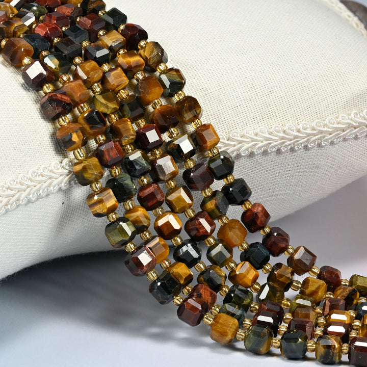 Tricolor tiger eye faceted cube beads 6mm with small gold spacer beads, several strands draped over a white cushion