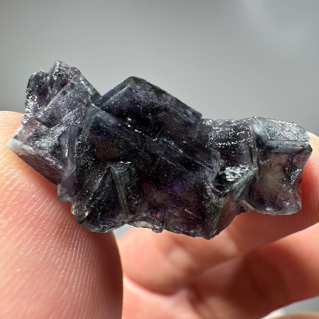 Dark purple cubic fluorite held between finger and thumb, stepped crystal faces catching the light