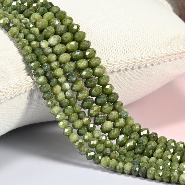 South Green Jade faceted rondelle beads draped over a cream fabric cushion, olive through deep forest green with fine dark speckling across the 4x6mm and 5x8mm strands.