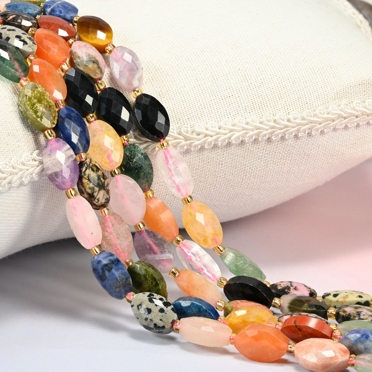 Assorted gemstone faceted flat oval beads 9x14mm with small gold seed bead spacers, several strands draped over a white cushion