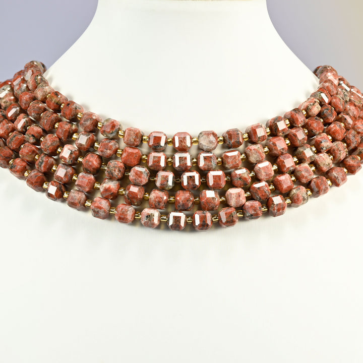 Red sesame jasper faceted cube strands arranged as a layered collar on a white display bust, brick red speckled beads across the neck
