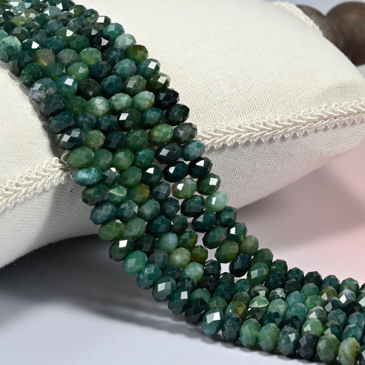 Moss agate faceted rondelle beads draped over a white fabric cushion with braided trim.
