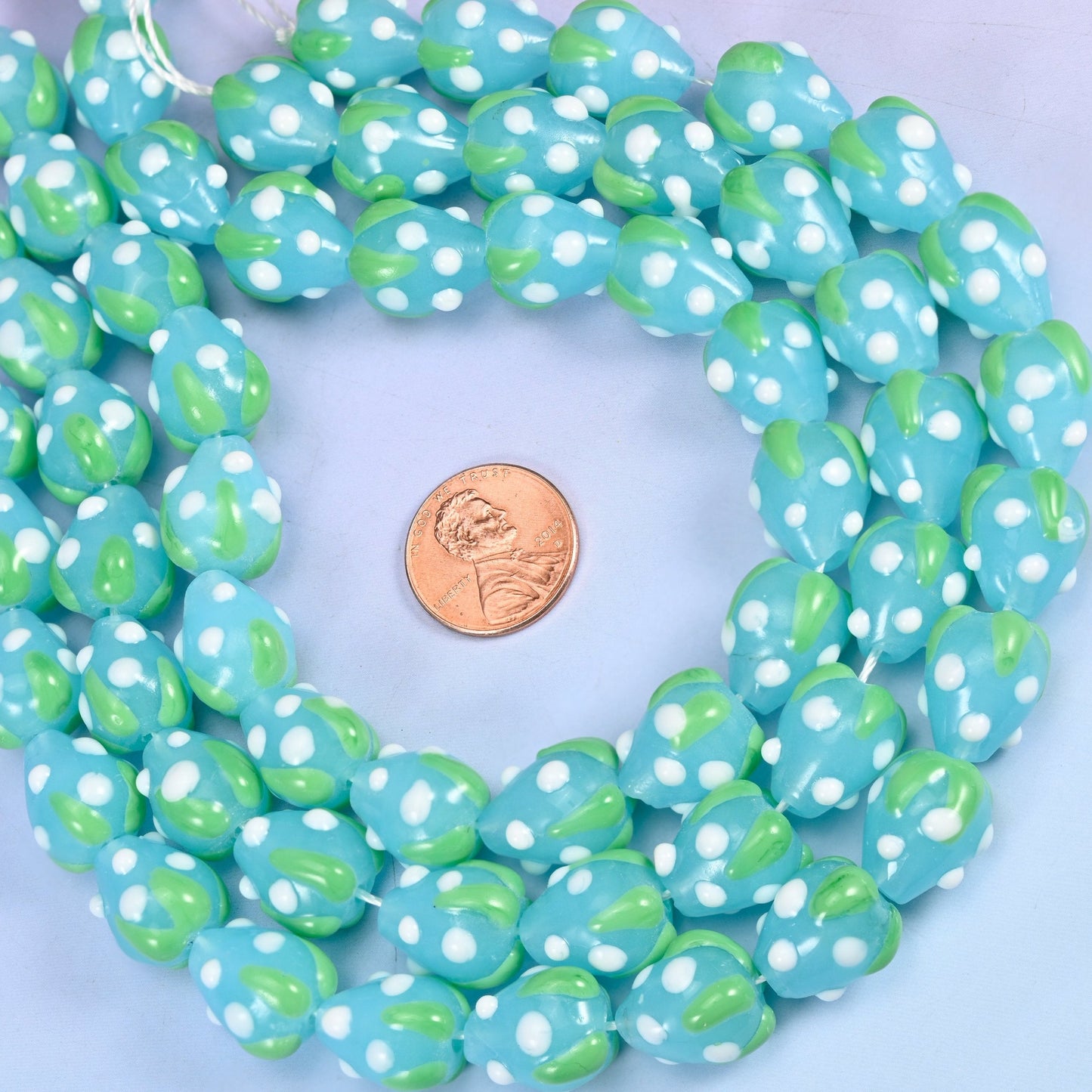 Sky blue opaque glass ovals scattered with small white seed dots and capped with an apple-green leafy band, so every bead reads as a little blue strawberry.