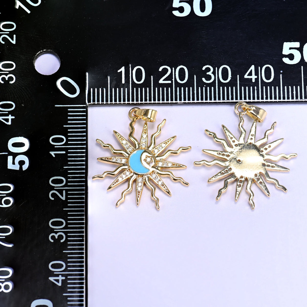 Enamel moon sun zirconia charms on a black millimetre ruler, one blue enamel pendant face up and one showing the gold reverse.