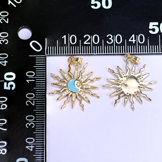 Enamel moon sun zirconia charms on a black millimetre ruler, one blue enamel pendant face up and one showing the gold reverse.