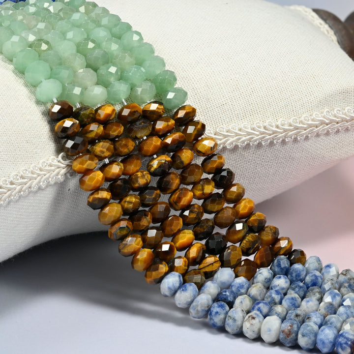 Chakra faceted rondelle beads 5x8mm draped over a white cushion, blocks of green aventurine, tiger eye and blue spot jasper