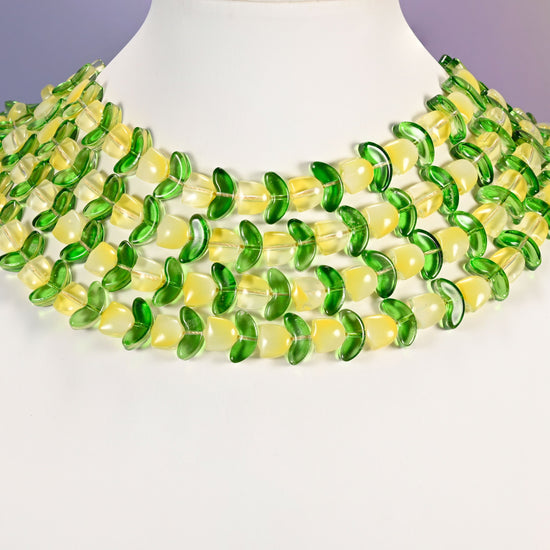 Multi-strand necklace of yellow tulip and green leaf glass beads laid on a white mannequin neck form against a lavender background.