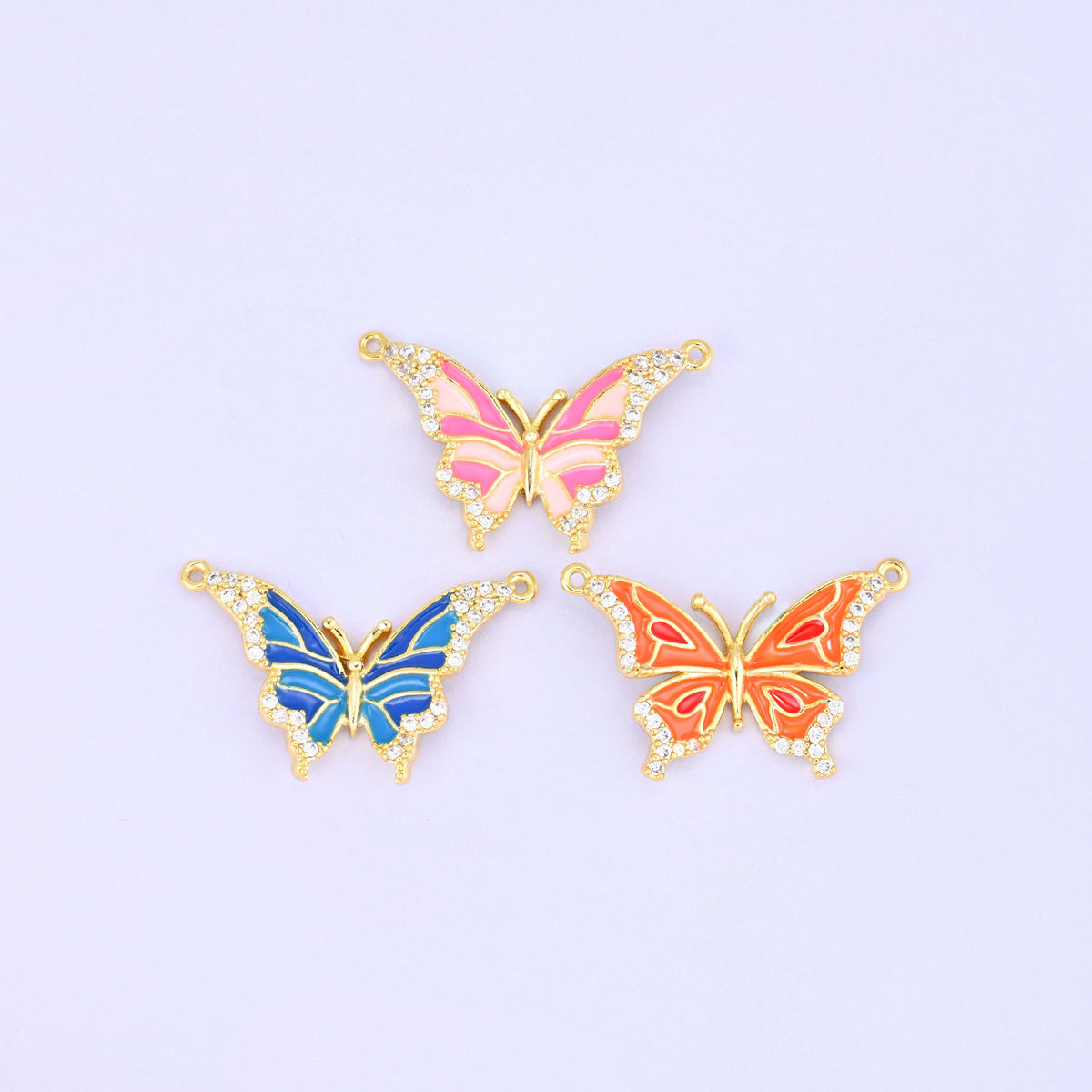 Three enamel butterfly double connector pendants shown flat and face up, the pink, blue and orange colourways together on a lavender background.