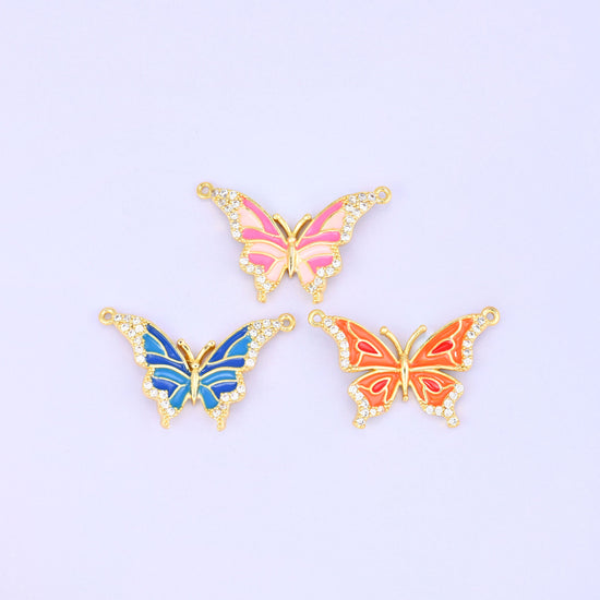 Three enamel butterfly double connector pendants shown flat and face up, the pink, blue and orange colourways together on a lavender background.