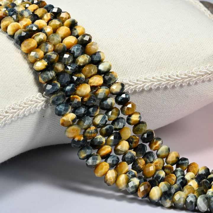 Dream Tiger Eye faceted rondelle beads draped over a cream fabric cushion, golden honey banded with steel blue-gray across the 4x6mm and 5x8mm strands.