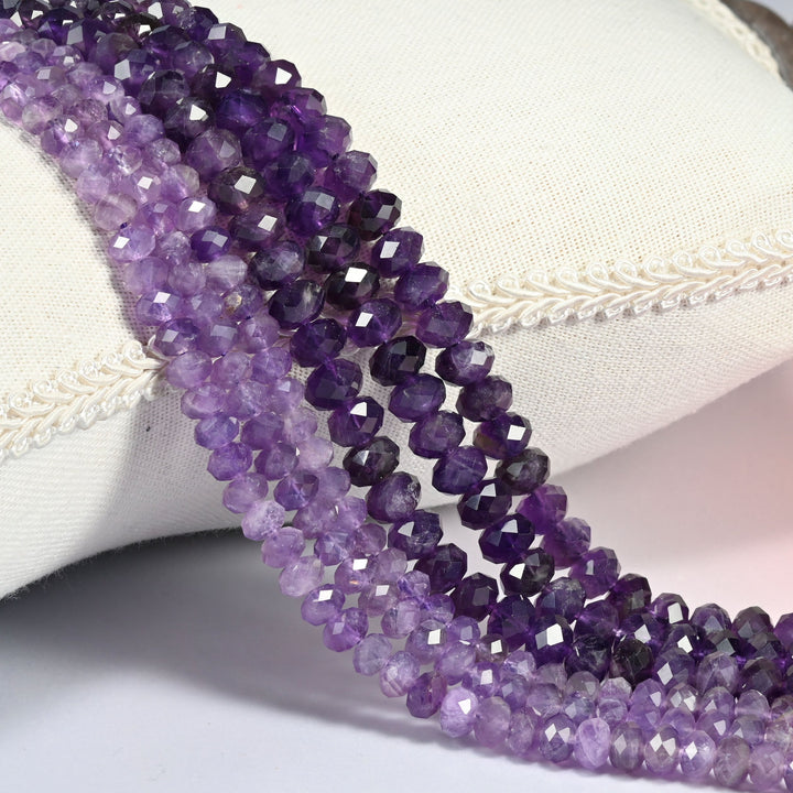 Purple amethyst faceted rondelle beads in translucent violet purple, three strands draped over a cream linen cushion with braided trim