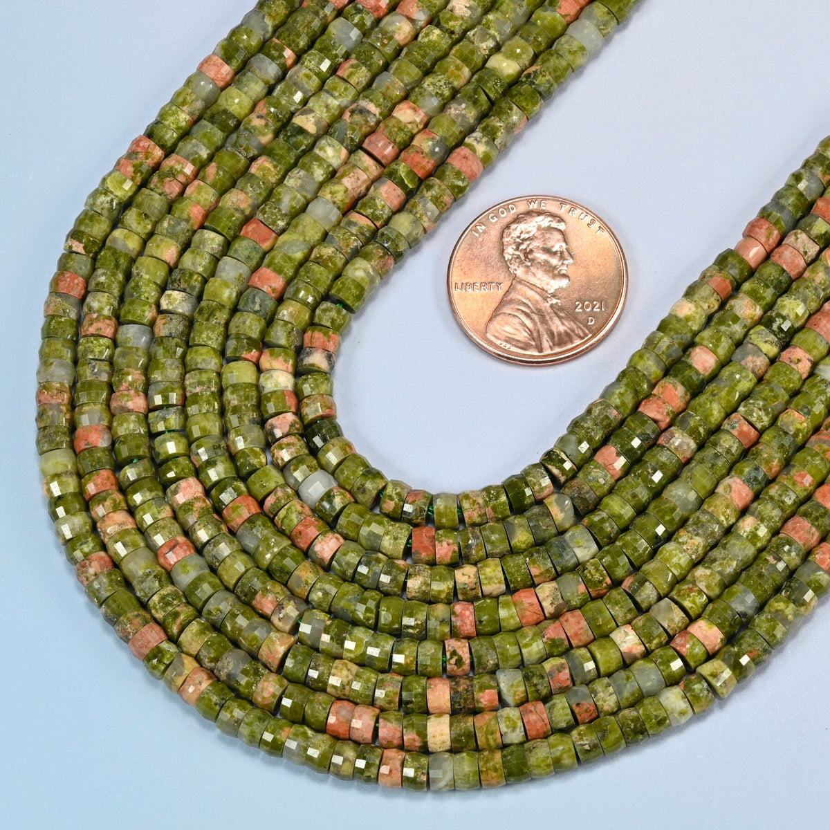 Close view of unakite faceted heishi disc strands curving beside a US penny for scale