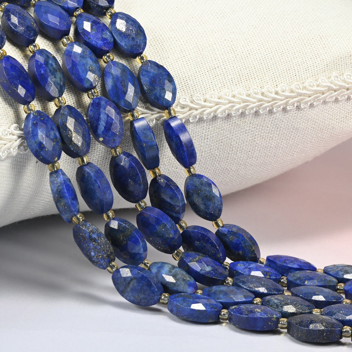 Lapis lazuli faceted flat oval beads 9x14mm with small gold seed bead spacers, several strands draped over a white cushion