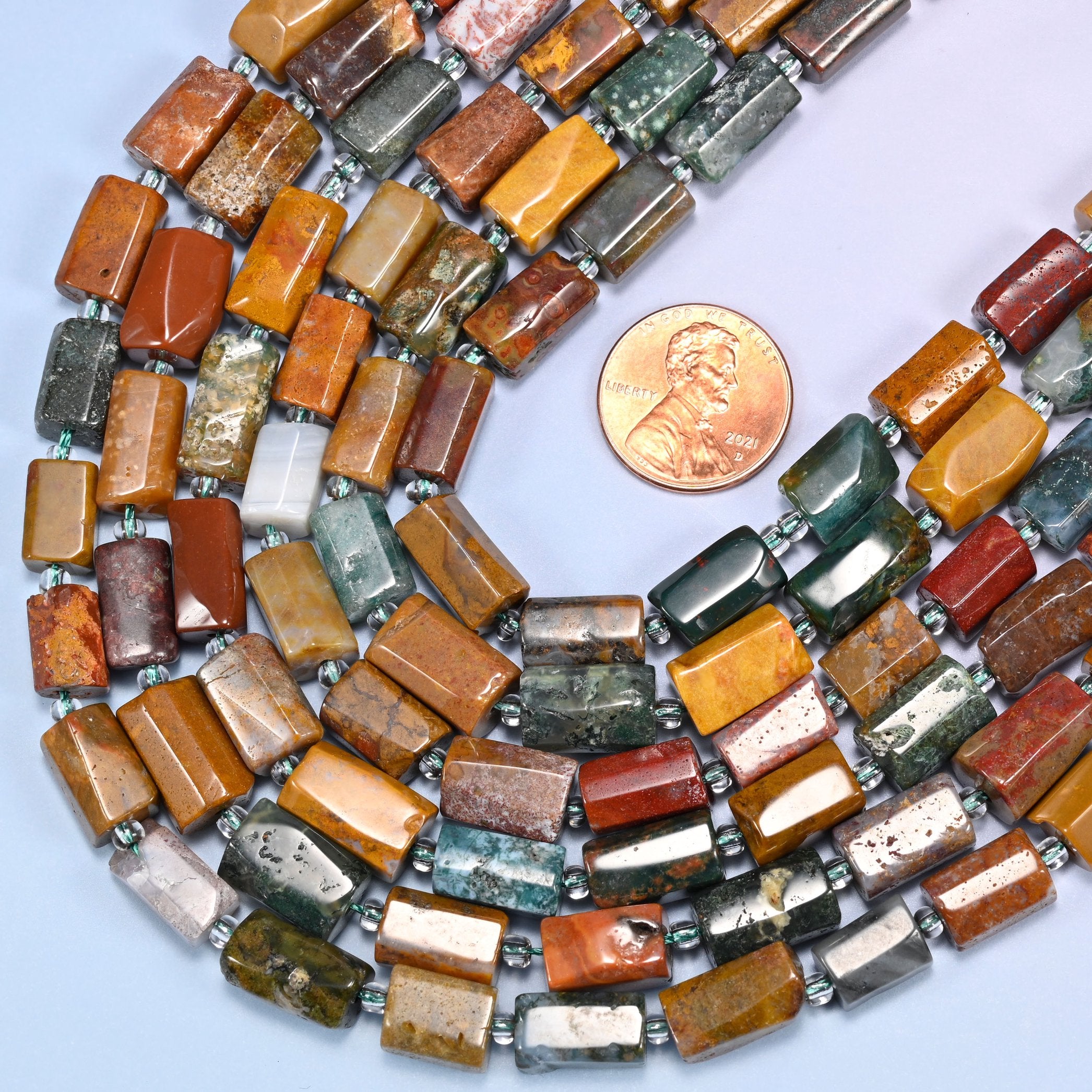 Rows of faceted Ocean Jasper tube beads laid straight on a lavender surface, showing the range of natural mustard, orange, and green coloring.