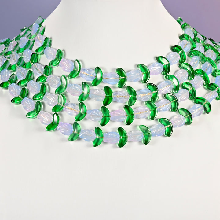 Multi-strand necklace of white tulip and green leaf glass beads laid on a white mannequin neck form against a lavender background.