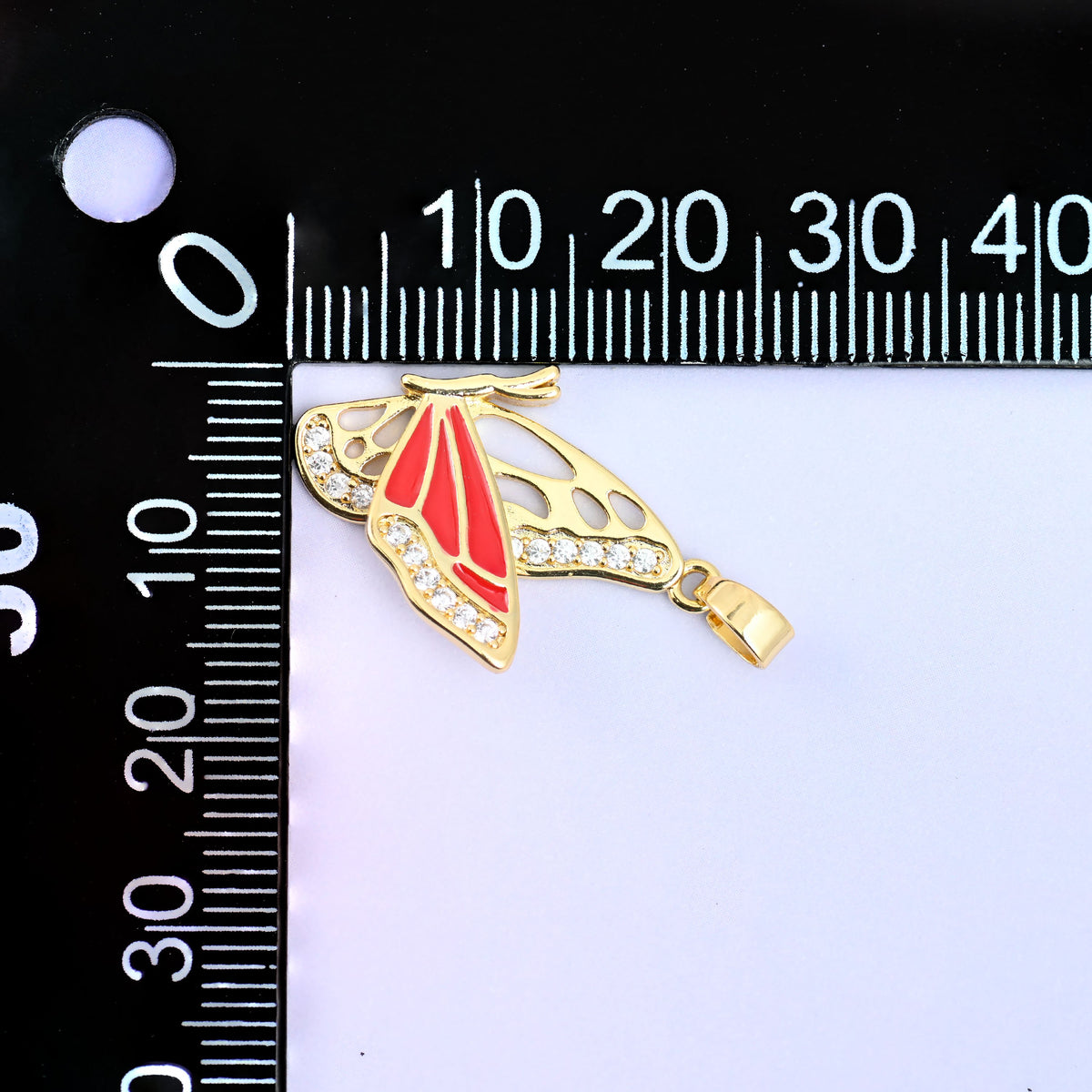 Flying butterfly charm pendant measured on a millimetre ruler, the red charm turned sideways with its bail to the right against the millimetre scale.