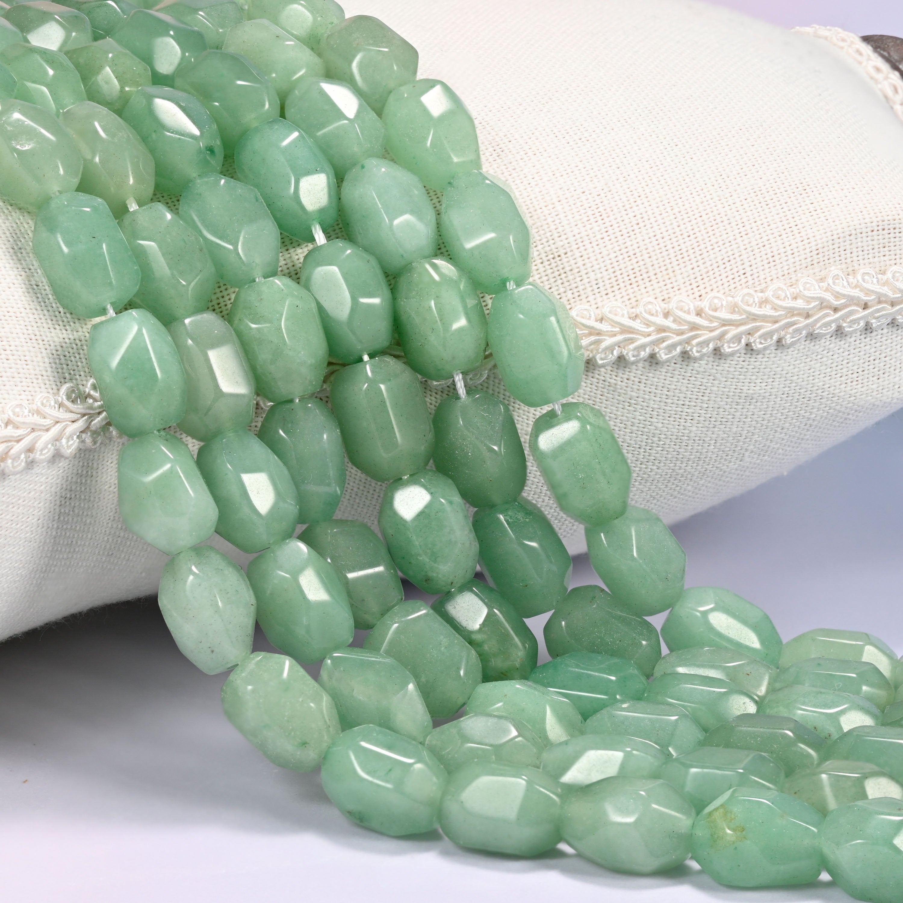 Close-up of multiple strands of green faceted Aventurine barrel beads cascading diagonally over a cream woven pillow with decorative trim.