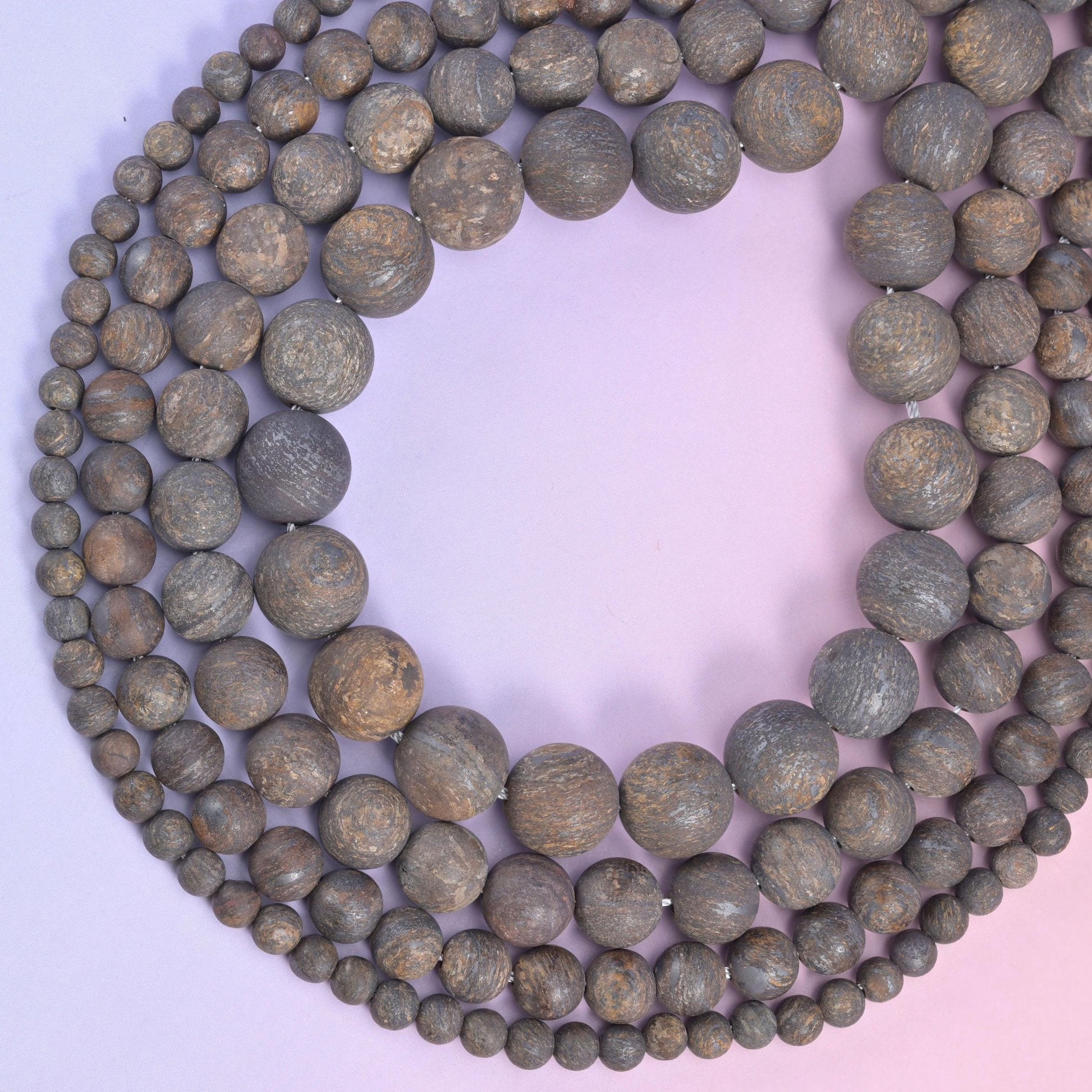 Open loop of mixed-size bronzite rounds on a lilac backdrop, drilled beads strung on cotton cord