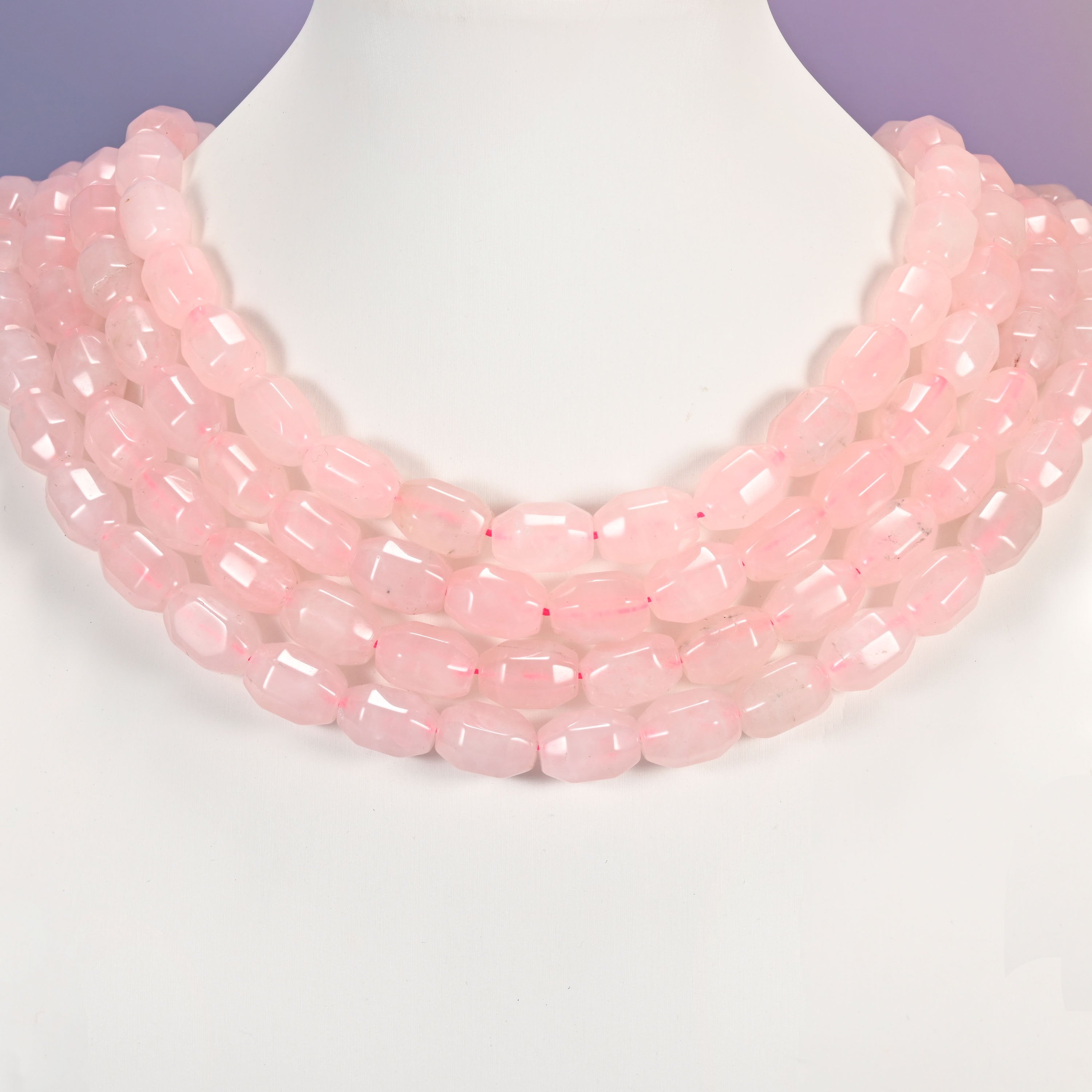 Multi-strand necklace of Rose Quartz faceted barrel beads laid on a white mannequin neck form against a lavender background.