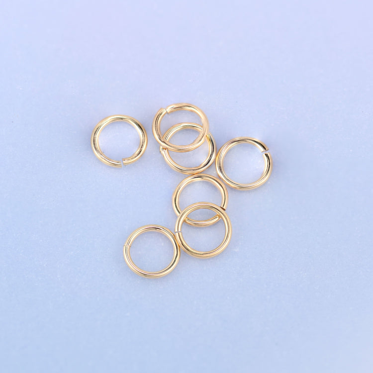 Seven 14K gold filled open jump rings grouped together, some lying flat and one standing on edge, showing the round wire profile and the small split in each ring.