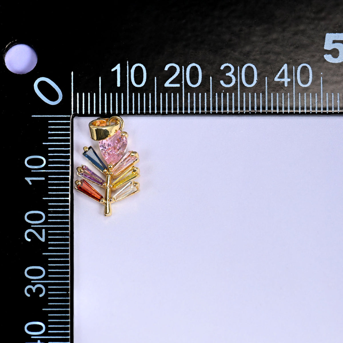 Multicolour leaf charm in gold with a pink stone and green, blue and orange baguette stones, beside a millimetre ruler for scale