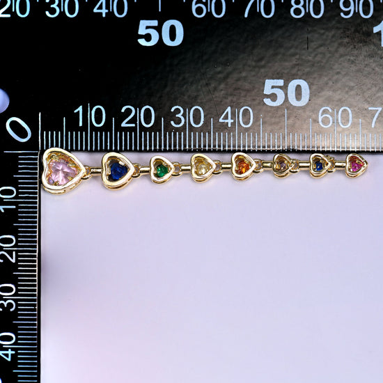 Pink heart pendant chain, a line of small gold-set heart stones in pink, blue, green and yellow, laid along a millimetre ruler for scale