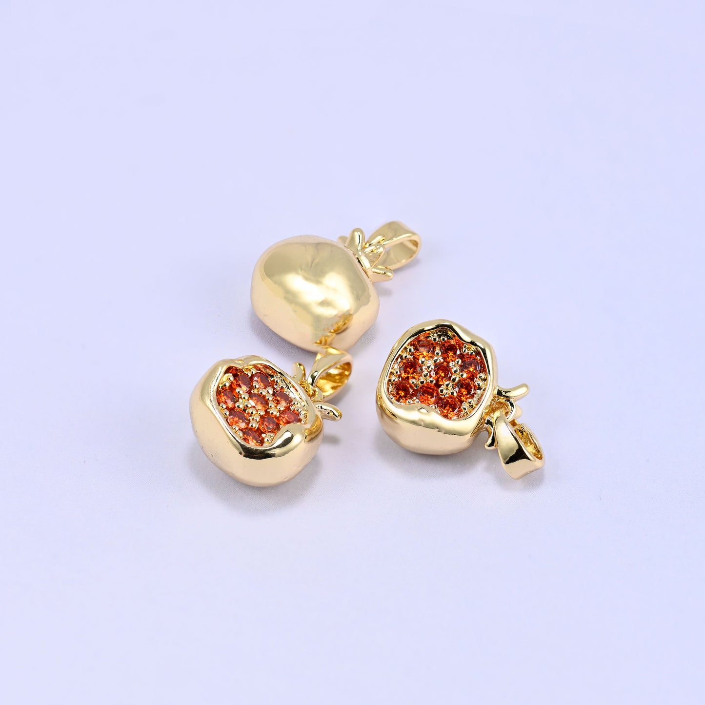 Three gold-plated pomegranate charms with orange zircon pave on a lilac background