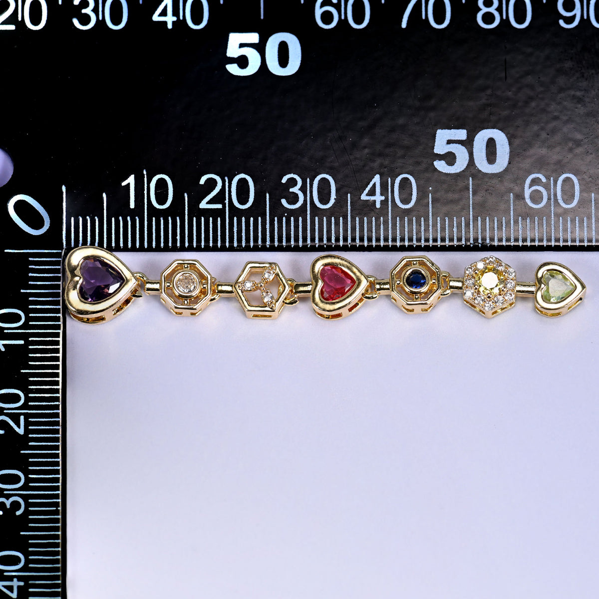 Purple heart pendant chain, a line of gold-set heart and round stones in purple, red, blue and clear, laid along a millimetre ruler for scale