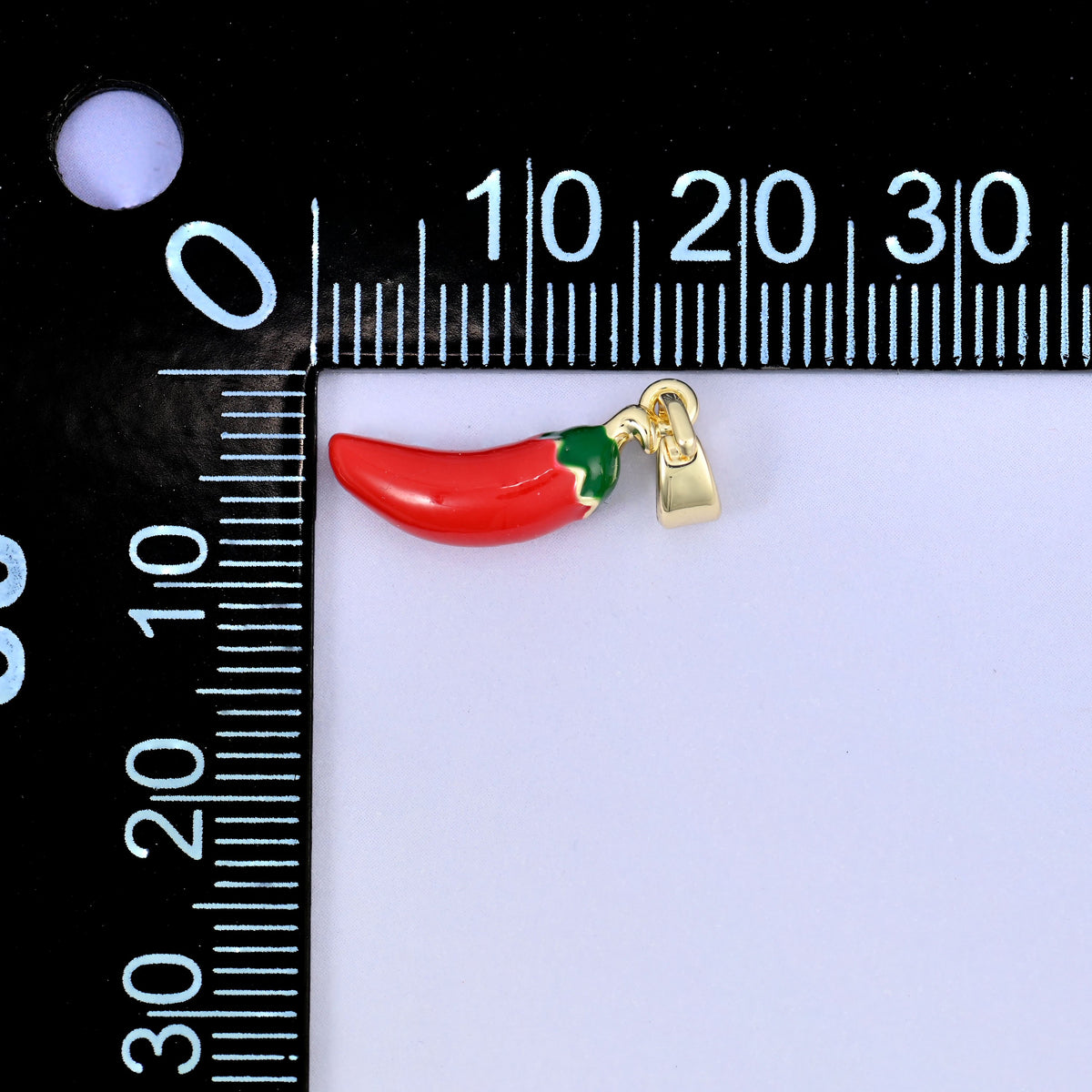 A red enamel chili pepper charm with a green stalk, lying sideways beside a millimetre ruler for scale