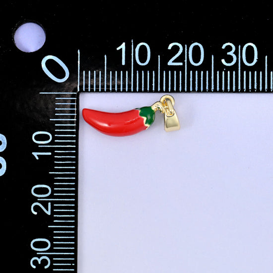 A red enamel chili pepper charm with a green stalk, lying sideways beside a millimetre ruler for scale