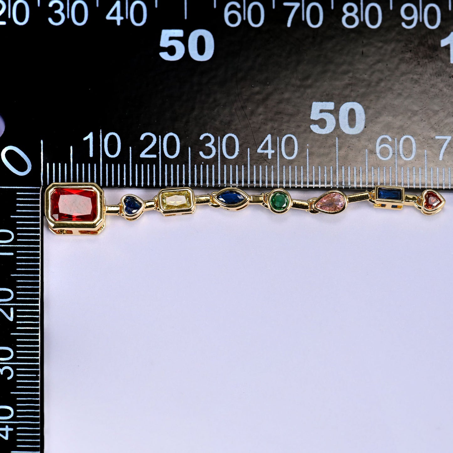 Red rectangle pendant with a chain of blue, green, pink and clear stones in gold, laid along a millimetre ruler for scale