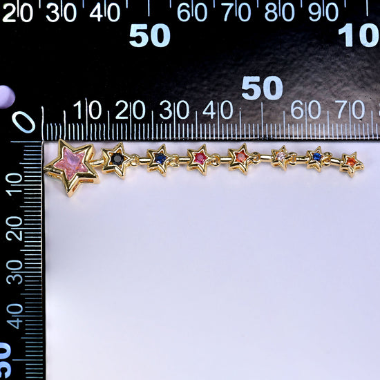 Shooting star pendant, a trail of small gold stars set with pink, blue and green stones leading to a larger pink star, laid along a millimetre ruler for scale