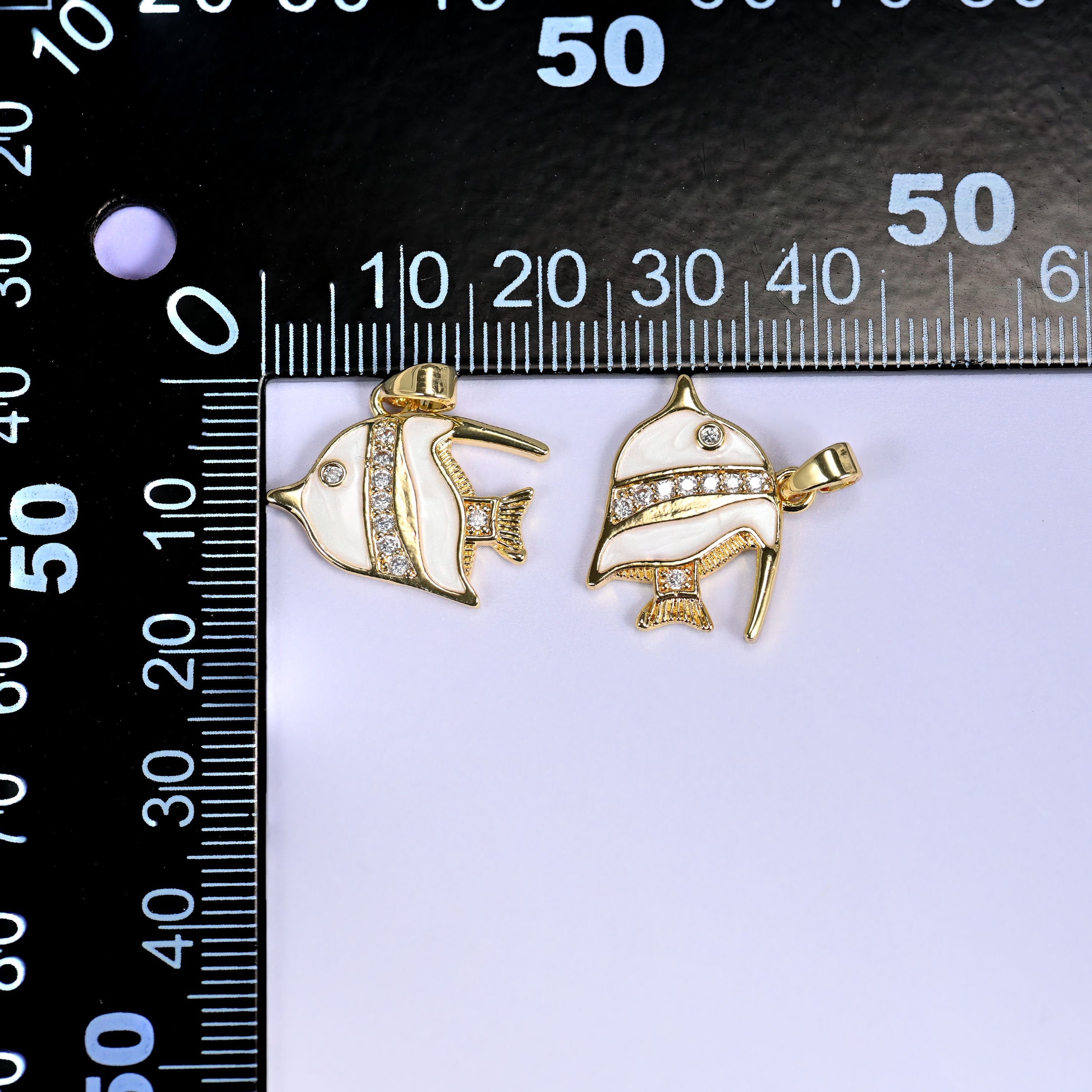 Two tropical fish charms in white enamel with gold fins and clear pave stones, shown front and angled, beside a millimetre ruler for scale